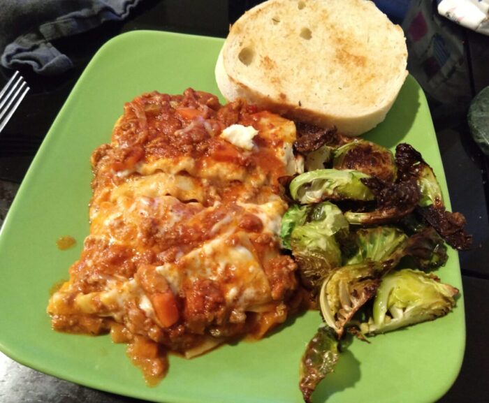 a plate of lasagna, roasted brussels sprouts, and a piece of garlic bread