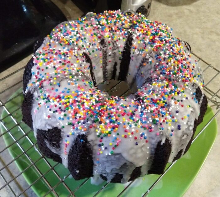 A darkly chocolate bundt cake topped with vanilla glaze and round rainbow sprinkles