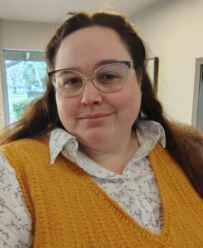 Me wearing a button-down shirt with a hand-knit yellow sweater vest. I have my hair down and I'm smiling at the camera.