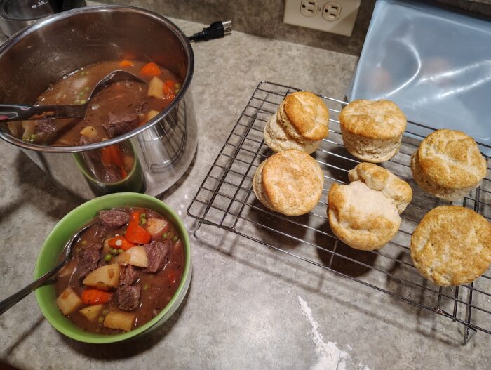 A bowl of stew, a big pot of stew, and biscuits cooling on a wire rack. All positioned near each other on the kitchen counter