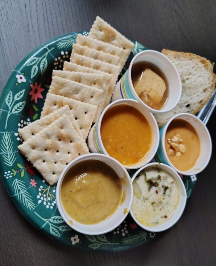 Five small cups of soup on a plate with crackers and bread