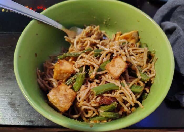 a bowl of soba noodles and green beans in peanut sauce along with chunks of baked tofu