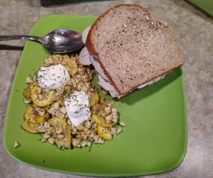 A sandwich next to a pile of roasted corn and summer squash