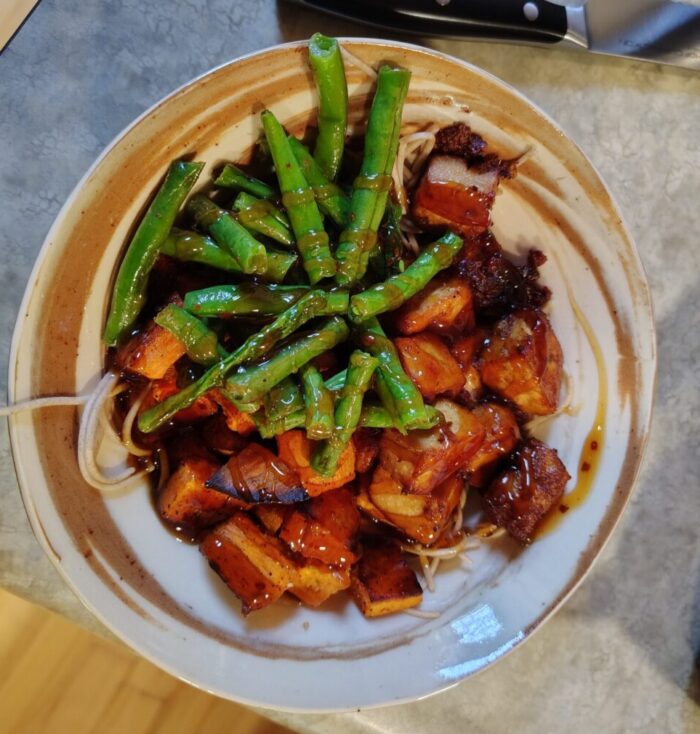 A bowl of soba noodles, roasted green beans, sweet potato, and baked tofu, drizzled with Panda express orange sauce