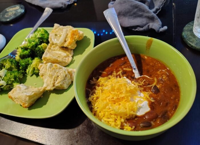 a bowl of bean chili topped with cheese and sour cream, next to a plate of broccoli and biscuits
