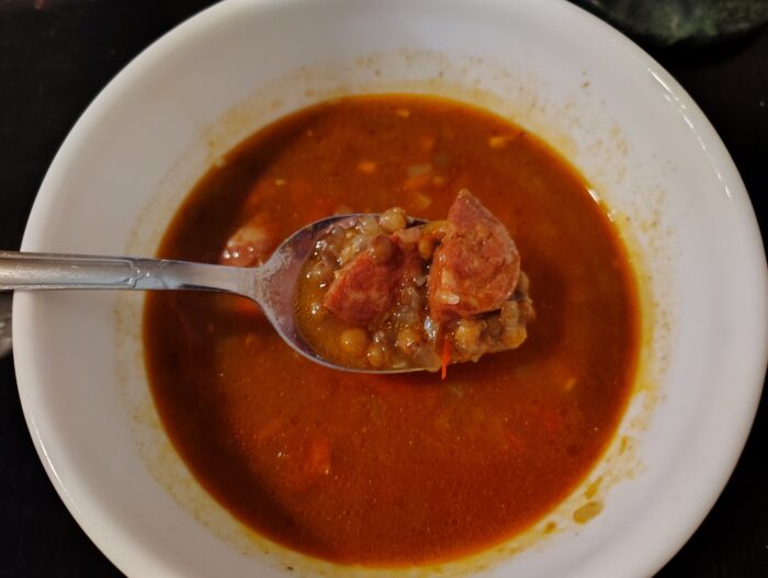 a bowl of soup with a spoon held above it showing a pile of lentils and sausage chunks