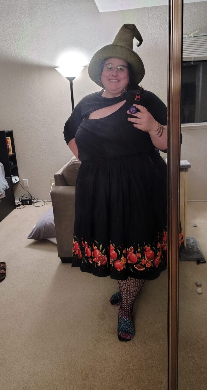 A mirror selfie of me in a black shirt with an aysmmetrical boob window, a black skirt with a pomegranate pattern along the bottom, black fishnets, and a mossy green witch hat