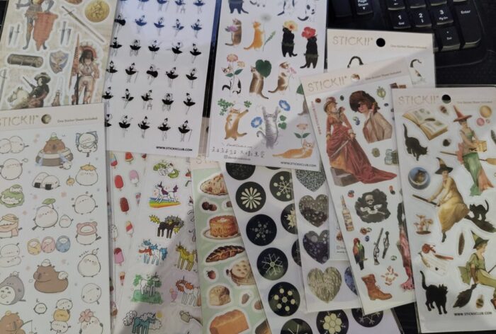 a pile of sticker sheets of many varieties including snowflakes, ladies with swords, capybaras, popsicles, unicorns, and cats with flowers