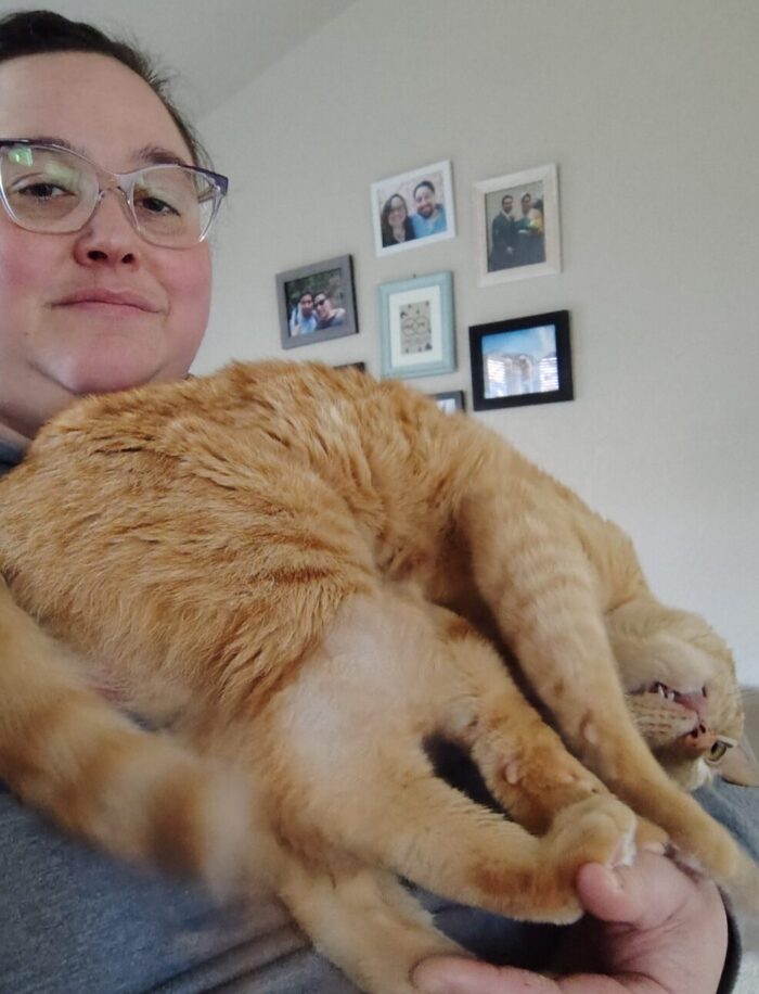 Fritz the cat leaning against my chest with all four paws in my hand. He has curled himself so his head is upsidedown and his tongue is sticking out. He looks fucking nuts