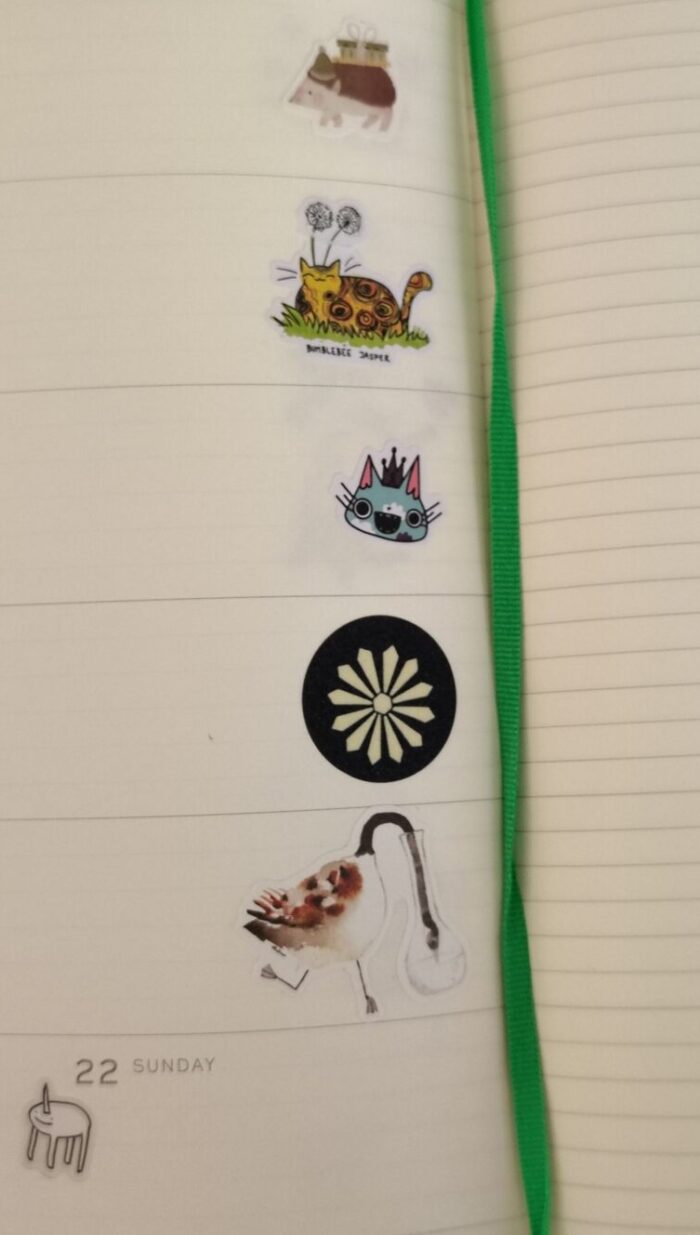 stickers in my planner including a hedgehog carrying gifts, a stylized cat face wearing a crown, a snowflake, a goose drinking from a case, and a ridiculous simplistically drawn unicorn