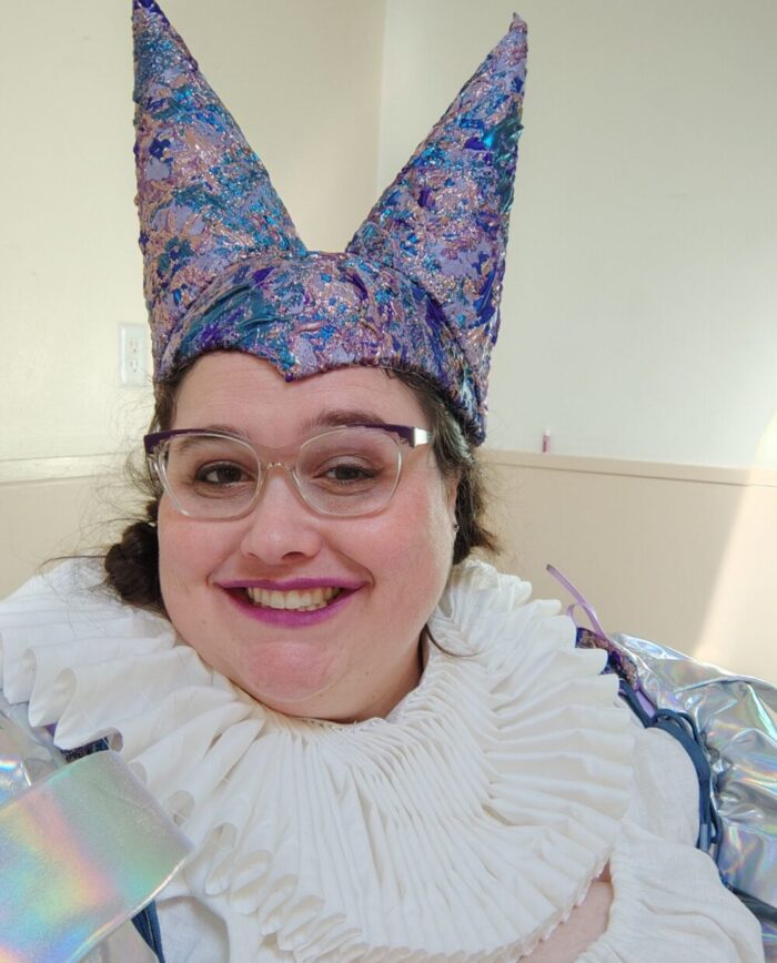 headshot of me in my jester outfit, featuring a purple and blue shiny double hennin and a white ruff