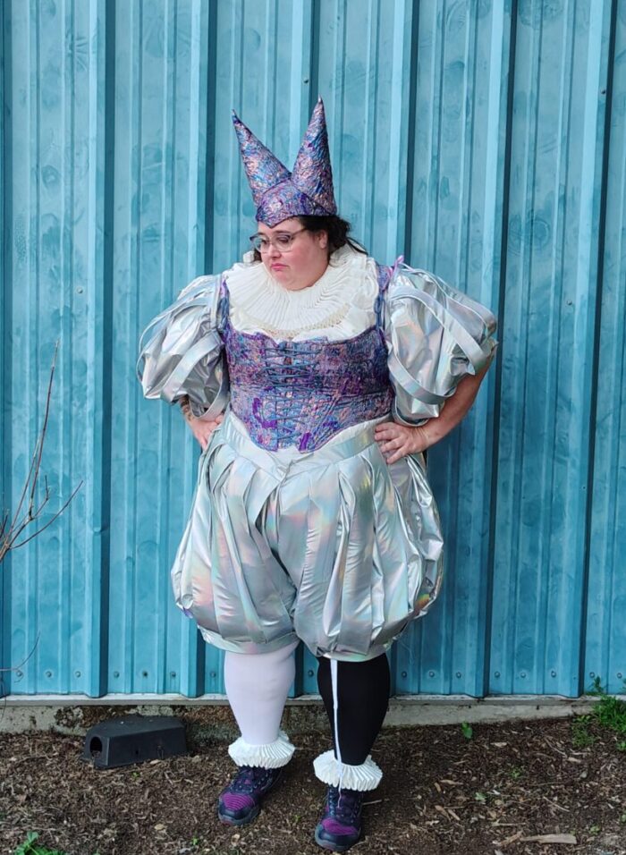 me, fully decked out in my jester outfit, momentarily forgetting to mask and going neutral/sad face, looking dejectedly at the ground, my hands on my hips