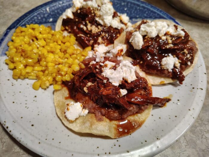 a plate with three tostadas topped with refried beans, pork in a chili sauce, and queso fresco. plus some corn!