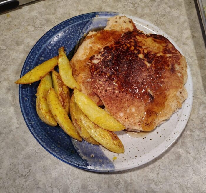A plate of buckwheat pancakes topped with maple syrup, and roasted potato wedges