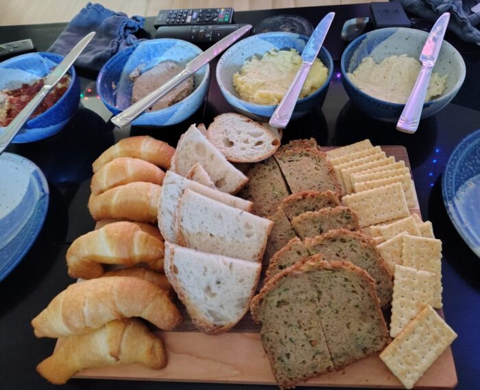a cutting board with crescent rolls, rosemary bread, zucchini bread, and club crackers next to bowls of compound butter