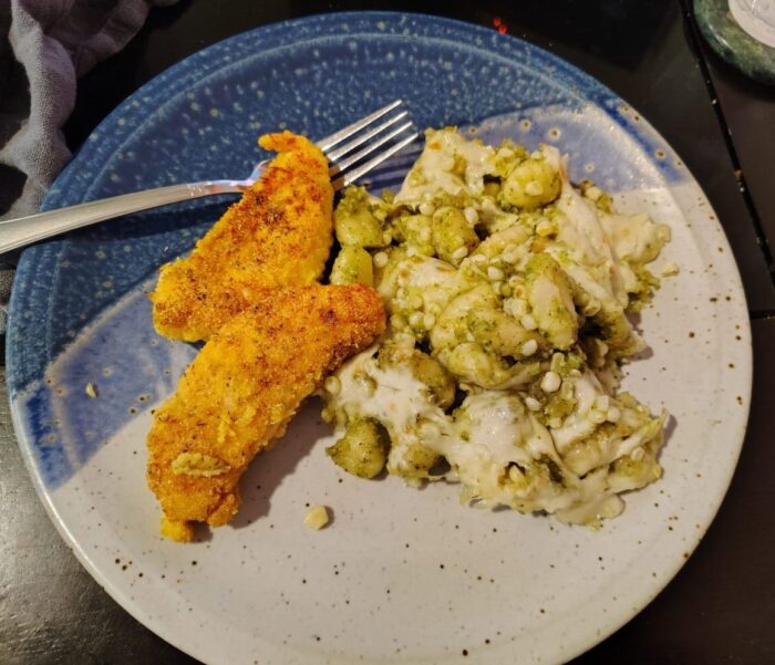 a plate of gnocchi with corn and pesto, topped in melted cheese. Two cornmeal-coated chicken tenders