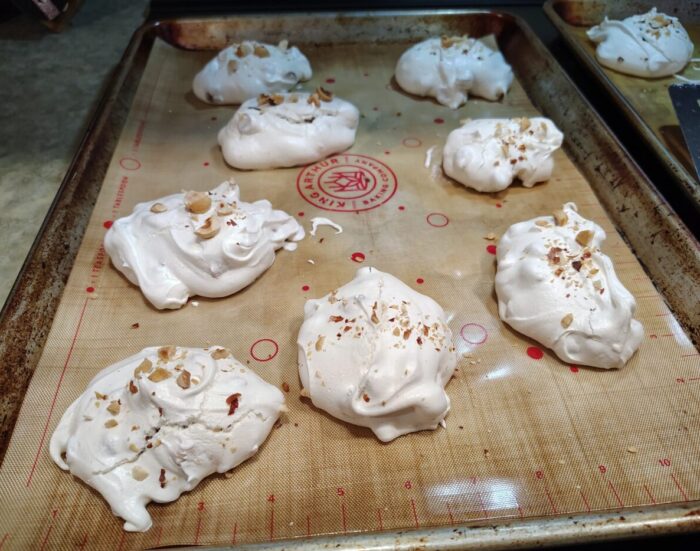 hazlenut and chocolate chip merengues on a sheet pan. They are topped with hazelnut bits. They are not shaped in any fancy way, just plopped onto the pan