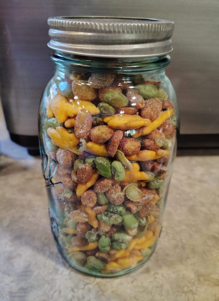 a glass jar of roasted edamame, honey roasted peanuts, cheese crackers, and spiced pecans