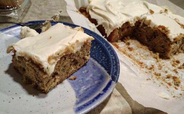 a zucchini sheet cake with cream cheese frosting. A slice of cake is on a plate with the remaining cake in the background