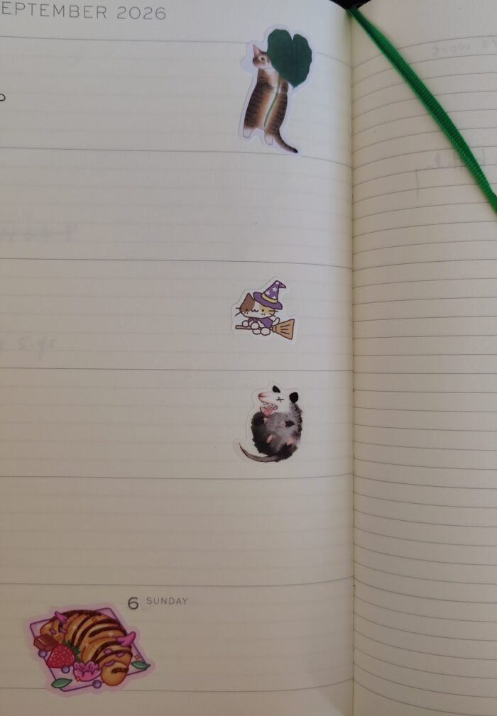 Stickers in my planner including a cat holding a leaf, a witch cat riding a broom, a screaming opossum, and a croissant